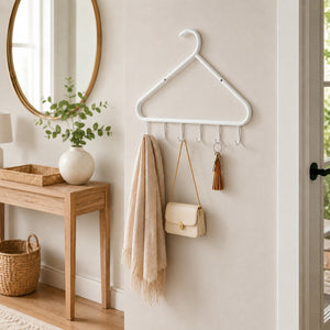 A white metal hanger coat rack with hooks mounted on a beige wall in a styled entryway. A cream scarf, a small cream handbag, and a tassel keychain hang from the rack.