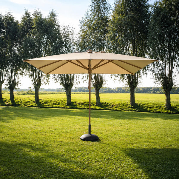 A large beige patio umbrella with a black weighted base stands on a green lawn in front of a row of tall trees under a bright sky.