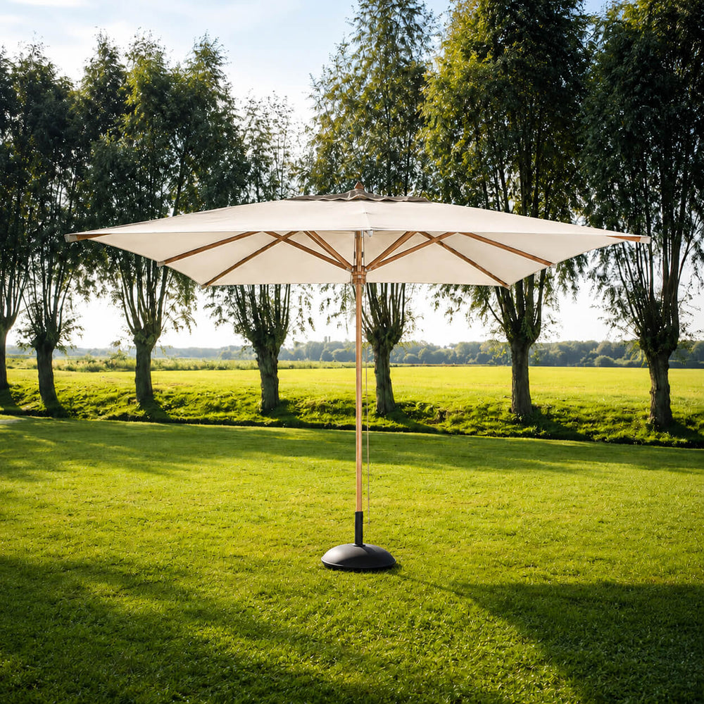 A large patio umbrella with a beige canopy and a bronze-finished central pole stands on a green lawn in front of a row of trees under a blue sky.