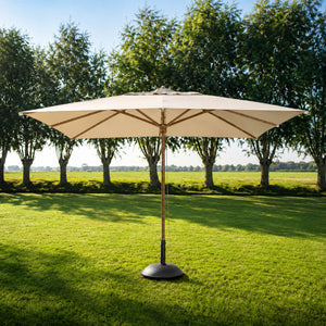 A large patio umbrella with a beige canopy and a bronze-finished central pole stands on a green lawn in front of a row of trees under a blue sky.