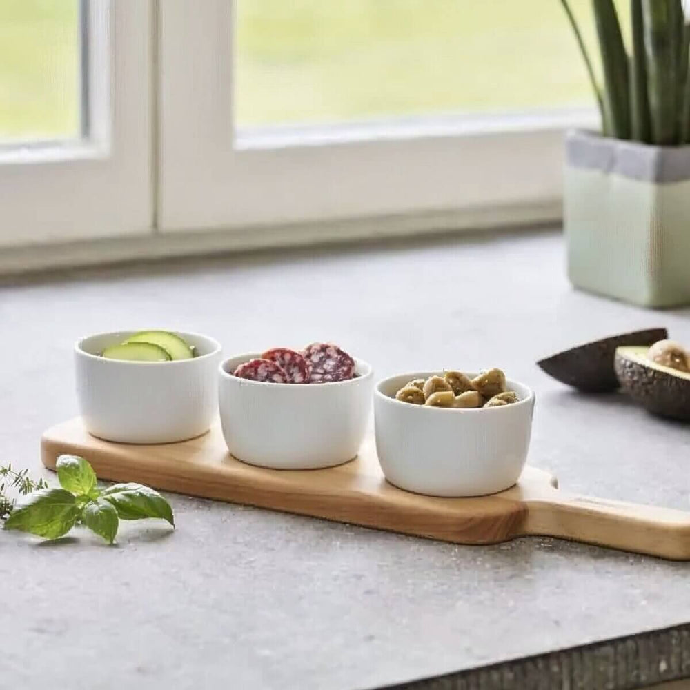 Three white bowls with snacks on a wooden tray on a kitchen counter.