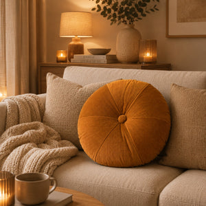 A round tufted mustard-orange throw pillow resting on a beige sofa in a warm, cozy living room setting with a knitted blanket, candles, and a lamp in the background.