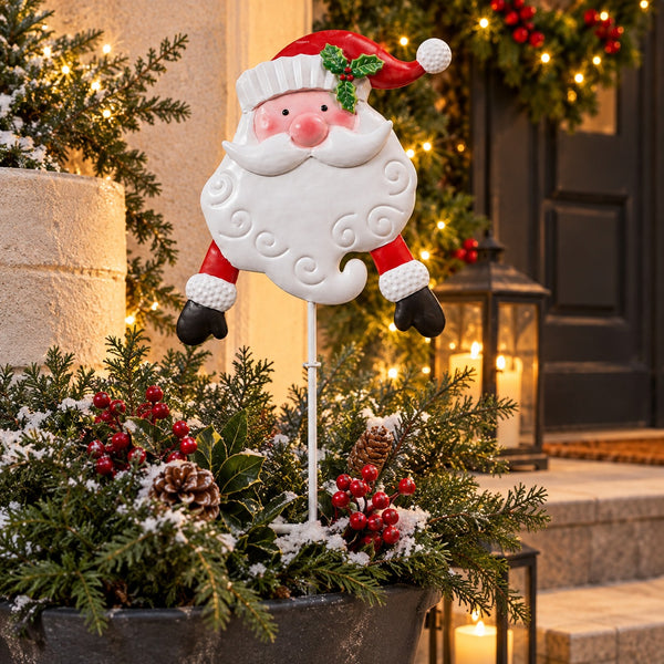 A decorative Santa Claus yard stake with a white beard, red hat, and black gloves stands in a holiday garden planter with evergreen branches, pinecones, and red berries. The background shows a warmly lit doorway with Christmas garlands and string lights.