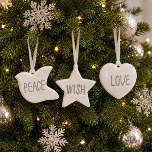 Three white ceramic Christmas tree ornaments shaped like a dove, star, and heart, hanging on a green Christmas tree with lights. The dove ornament is written with the word 'PEACE', the star with 'WISH', and the heart with 'LOVE'.