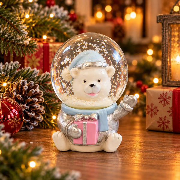 A Christmas-themed snow globe figurine featuring a white polar bear wearing a light blue hat and scarf, holding a pink gift box with silver trim. The figurine is set on a wooden surface surrounded by festive decorations, including pine branches, a pinecone, and warm bokeh lights in the background.