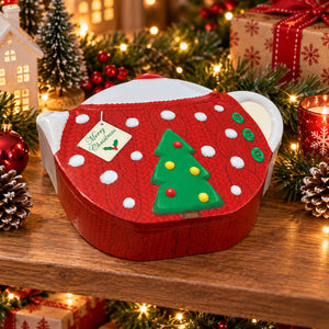 A red Christmas-themed gift box with a white lid, decorated like a knitted sweater with white polka dots, a green Christmas tree, and a 'Merry Christmas' tag, displayed on a wooden shelf with festive lights and ornaments.