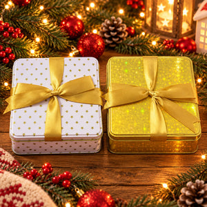 Two square metal gift boxes tied with wide gold ribbons, placed on a wooden surface and surrounded by Christmas decorations including pine branches, red ornaments, and warm string lights.