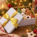 A white rectangular metal gift tin filled with assorted Christmas cookies, shown with the lid leaning against it and tied with a wide gold ribbon. The tin has small gold stars and sits on a wooden surface surrounded by holiday greenery, ornaments, and warm string lights.