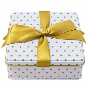A square white gift tin wrapped in a white paper pattern of small gold stars, tied with a wide gold satin ribbon in a bow, on a plain white background.