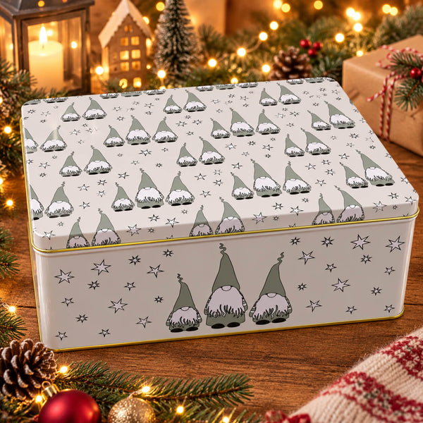 A rectangular metal gift tin with a lid, decorated with a repeating pattern of green and white gnomes and small stars on a warm off-white background. The tin is styled on a wooden surface with festive Christmas decorations, including pine branches, a lit candle, and warm bokeh lights in the background.