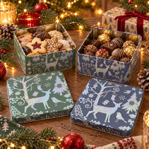 Three decorative square metal gift boxes filled with assorted cookies and chocolates, arranged on a wooden table with holiday lights, pine branches, and ornaments. The boxes and their lids feature white reindeer and snowflake designs in green, blue, and navy tones.