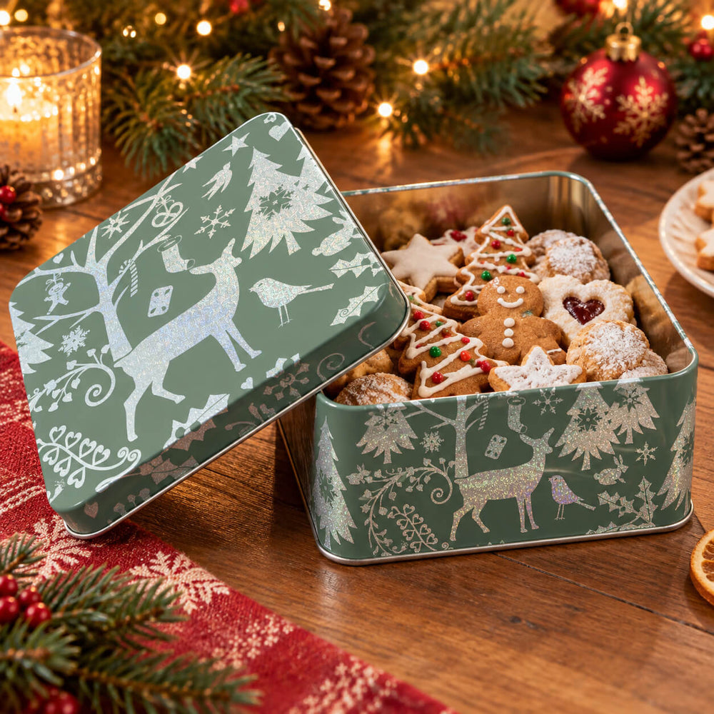 Three decorative square metal gift boxes filled with assorted cookies and chocolates, arranged on a wooden table with holiday lights, pine branches, and ornaments. The boxes and their lids feature white reindeer and snowflake designs in green, blue, and navy tones.