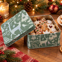 A square metal tin of holiday cookies sits open on a wooden table, with the lid leaning against it. The tin is sage green with white Christmas illustrations, including reindeer, trees, birds, and snowflakes.