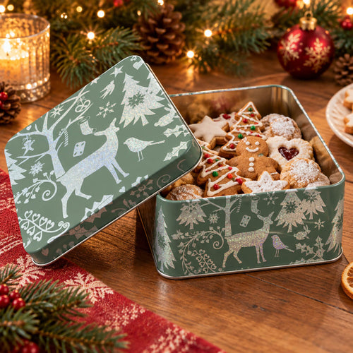 A square metal tin of holiday cookies sits open on a wooden table, with the lid leaning against it. The tin is sage green with white Christmas illustrations, including reindeer, trees, birds, and snowflakes.