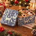 A square metal tin of holiday cookies sits open on a wooden table, with its lid leaning beside it. The tin is decorated with a winter scene in muted blue-gray and cream, featuring reindeer, trees, birds, and snowflakes.