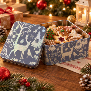 A square metal tin of holiday cookies sits open on a wooden table, with its lid leaning beside it. The tin is decorated with a winter scene in muted blue-gray and cream, featuring reindeer, trees, birds, and snowflakes.