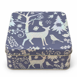 A square metal tin with rounded corners, decorated with a winter holiday pattern in white and iridescent silver. The design features silhouettes of a deer, birds, Christmas trees, snowflakes, and ornaments on a dark navy blue background.