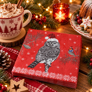 A stack of red Christmas paper napkins featuring a black-and-white owl wearing a Santa hat, surrounded by holiday decorations including a mug of whipped cream, pine branches, candles, and cookies on a wooden table.