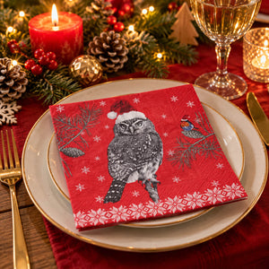 A festive Christmas-themed paper napkin featuring a gray and white owl wearing a red Santa hat, set on a gold-rimmed white plate in a holiday table setting with candles, pinecones, and a wine glass.