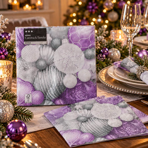 A boxed and folded paper napkin set from Cucina & Tavola is displayed on a decorated holiday table. The napkin packaging features a purple and gray Christmas ornament design, and the folded napkin shows a matching purple and gray pattern with sandwich cookies and ornaments.