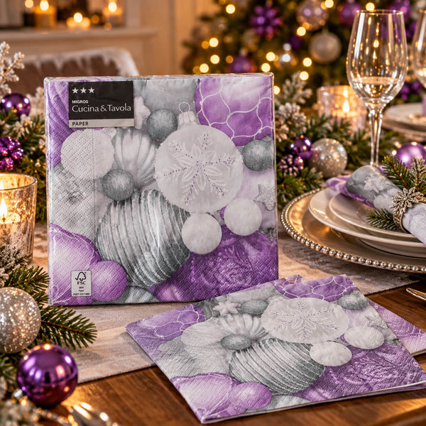 A boxed and folded paper napkin set from Cucina & Tavola is displayed on a decorated holiday table. The napkin packaging features a purple and gray Christmas ornament design, and the folded napkin shows a matching purple and gray pattern with sandwich cookies and ornaments.