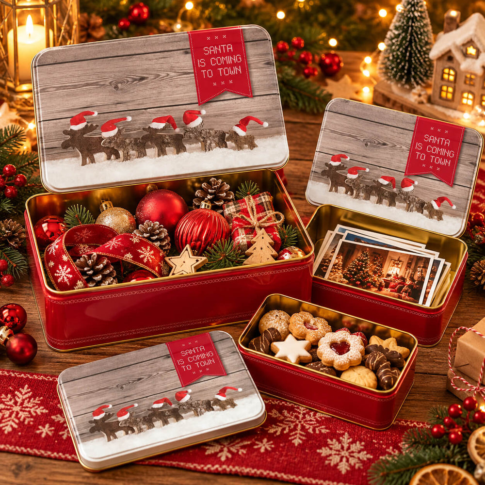 Three red Christmas-themed metal gift boxes with hinged lids are arranged on a decorated tabletop. The lids show a rustic wood background with a line of Santa-clad reindeer and a red tab reading “Santa Is Coming To Town.