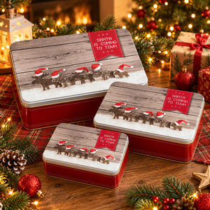 Three red metal gift boxes with wooden-look lids, each printed with Santa and reindeer carrying gifts and a red label reading "Santa is coming to town," arranged on a holiday table with Christmas lights, ornaments, pine branches, and wrapped presents.