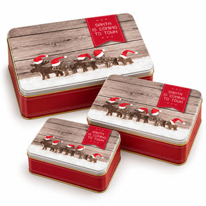 Three rectangular Christmas-themed metal gift boxes arranged in a pyramid on a white background. The boxes have red sides, gold trim, and lids featuring a rustic wood-plank design with a snowy scene, reindeer, and Santa hats, plus a red label that reads 'Santa is Coming to Town.