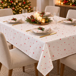 A white tablecloth with a red star pattern is draped over a dining table set for four with plates, wine glasses, gold cutlery, and a holiday centerpiece. A decorated Christmas tree and warm ambient lights are visible in the background.