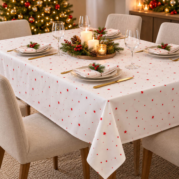 A white tablecloth with a red star pattern is draped over a dining table set for four with plates, wine glasses, gold cutlery, and a holiday centerpiece. A decorated Christmas tree and warm ambient lights are visible in the background.
