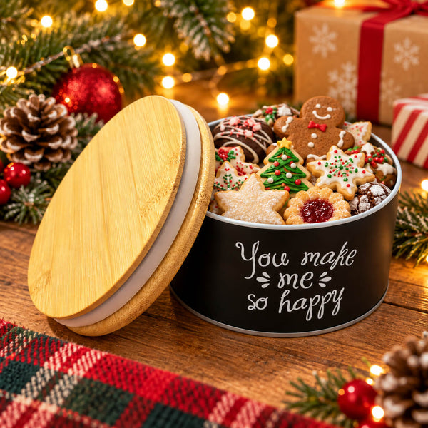 A round black tin filled with assorted decorated cookies and a large bamboo-and-white sandwich cookie on the lid, set on a wooden table with Christmas lights, pinecones, ornaments, and wrapped gifts in the background. The tin is printed with the phrase 'You make me so happy'.
