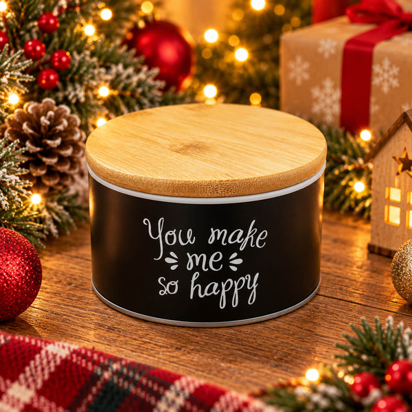 A cylindrical black tin with a light wood lid, featuring the phrase 'You make me so happy' in white script, set on a wooden table surrounded by Christmas decorations including lights, pinecones, ornaments, and wrapped gifts.