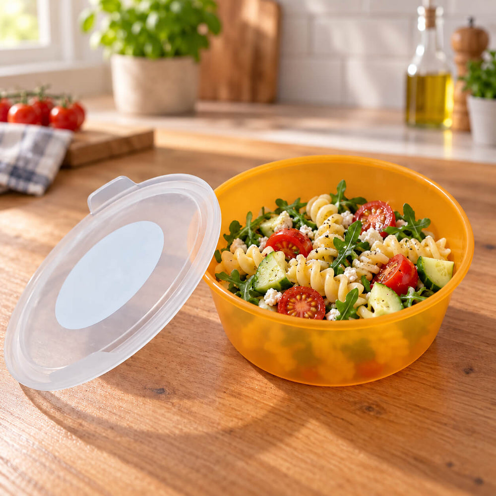 A translucent orange plastic food storage container with a matching translucent lid, shown open and filled with a pasta salad on a wooden countertop in a bright kitchen setting.