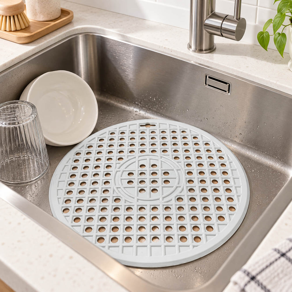 A round gray silicone kitchen mat with a textured grid pattern, placed inside a white farmhouse-style kitchen sink.