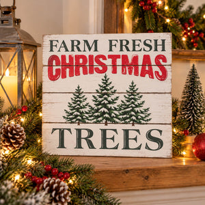 A rustic white wooden sign reading 'FARM FRESH CHRISTMAS TREES' with three green pine tree illustrations, displayed on a wooden mantel surrounded by holiday greenery, lights, pinecones, and a lantern.