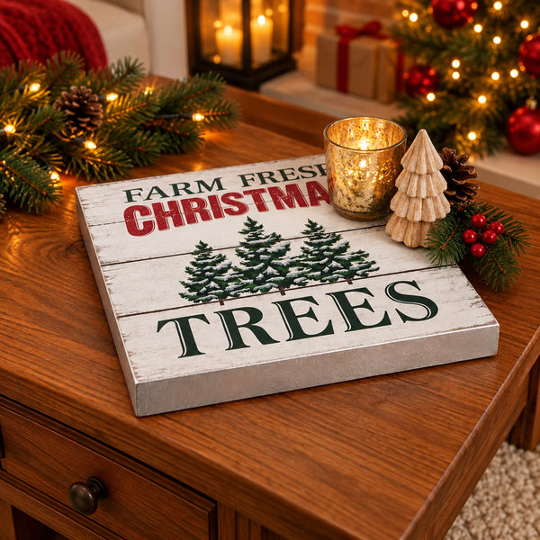 A square decorative sign with a whitewashed wood look reading “FARM FRESH CHRISTMAS TREES,” featuring three green pine tree illustrations. The sign is styled on a wooden table with holiday decorations including a gold candle holder, a small wooden tree, pinecones, and evergreen branches with warm lights in the background.