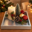 A silver rectangular tray holding a lit white pillar candle tied with twine, a small white pine tree, pinecones, greenery, and red and gold holiday ornaments on a wooden table.