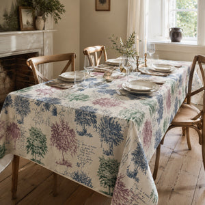 A rectangular dining table covered with a cream tablecloth featuring a vintage-style botanical tree pattern in muted blue, green, and purple. The table is set with white plates, clear wine glasses, and small floral centerpieces, surrounded by wooden dining chairs in a bright, traditional dining room with a fireplace and window.