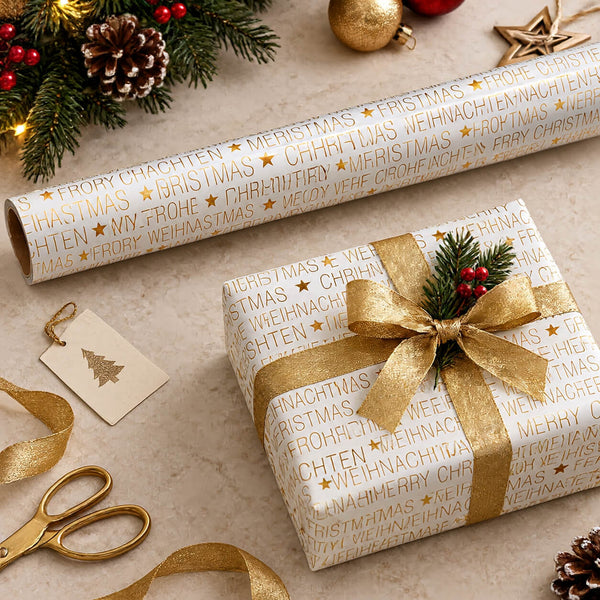 Gold and white Christmas wrapping paper with repeating festive text and stars, shown wrapped around a gift box and a roll on a styled tabletop with gold ribbon, scissors, pine branches, ornaments, and a kraft gift tag.