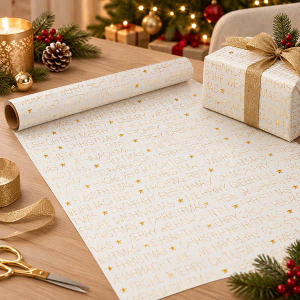 White wrapping paper with gold foil Christmas greetings and stars, shown partially unrolled on a wooden table with a wrapped gift box, ribbon, scissors, and holiday decorations in the background.