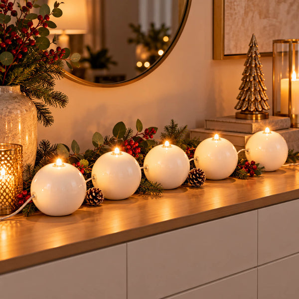 Five white spherical tea light candles lit in a row on a wooden console table, decorated with pinecones, evergreen branches, and red berries in a warm holiday interior setting.