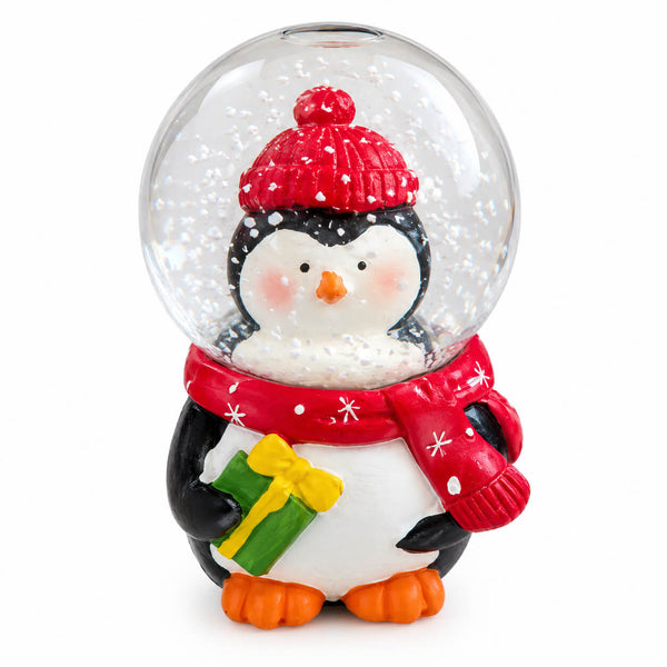 A penguin snow globe figurine wearing a red knit hat and matching red scarf, with orange feet, holding a green gift box with a yellow bow inside a clear round globe on a white background.