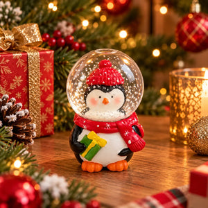 A festive penguin snow globe figurine wearing a red knit hat and scarf, holding a small green gift box, displayed on a wooden table with Christmas decorations including a wrapped gift, pinecones, ornaments, and warm string lights in the background.