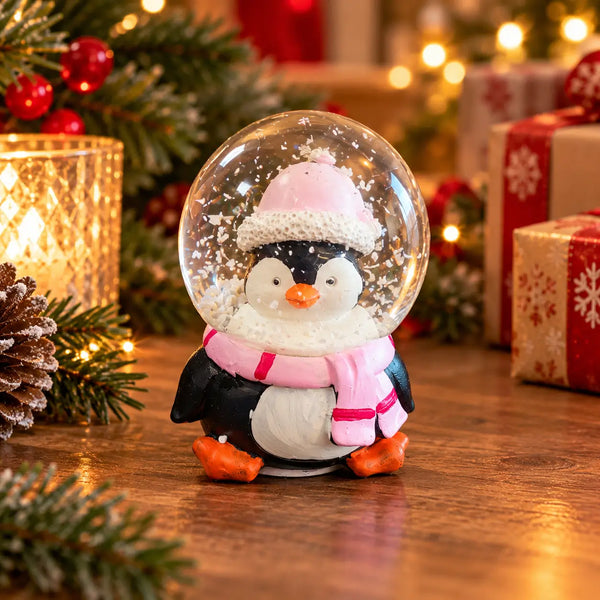 A penguin snow globe decoration on a wooden table, surrounded by a warm, festive Christmas background with pine branches, a lit candle, and wrapped gifts.