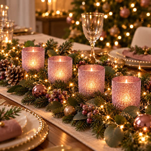 Four lit glittery pink cylindrical candle holders arranged on a decorated Christmas table runner with greenery, pinecones, berries, and warm string lights.