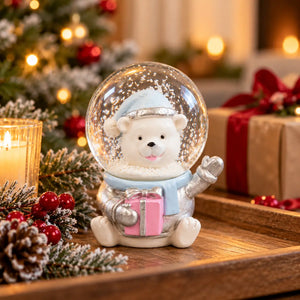 A Christmas-themed snow globe figurine featuring a white polar bear wearing a light blue hat and scarf, holding a pink gift box inside a clear globe. The figurine is styled on a wooden tray with a lit candle, pinecones, red berries, and wrapped presents in the background.