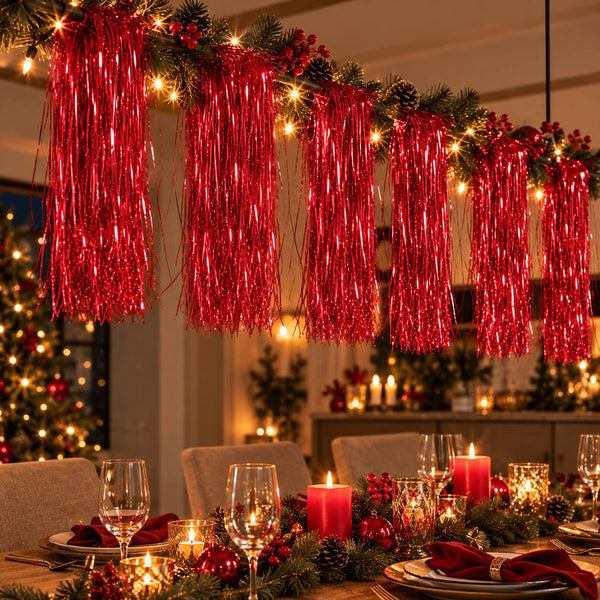 Red metallic tinsel fringe garland hanging across a decorated dining table with candles, plates, and holiday greenery in a warmly lit indoor setting.