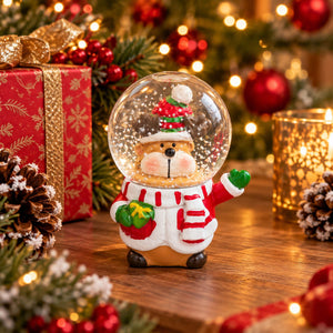 A festive Christmas snow globe figurine featuring a dog dressed as Santa Claus, standing on a wooden table with a blurred holiday background including a red gift box, pinecones, lights, and ornaments.