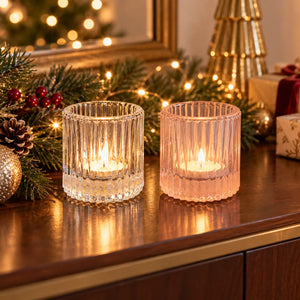 Two ribbed glass candle holders on a wooden table, one clear and one pale pink, each holding a lit tea light candle. The scene is decorated for Christmas with pine branches, lights, and ornaments in the background.