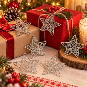 Five silver wire star-shaped ornaments arranged on a festive Christmas table with wrapped gifts, pine branches, red berries, and a lit candle in the background.
